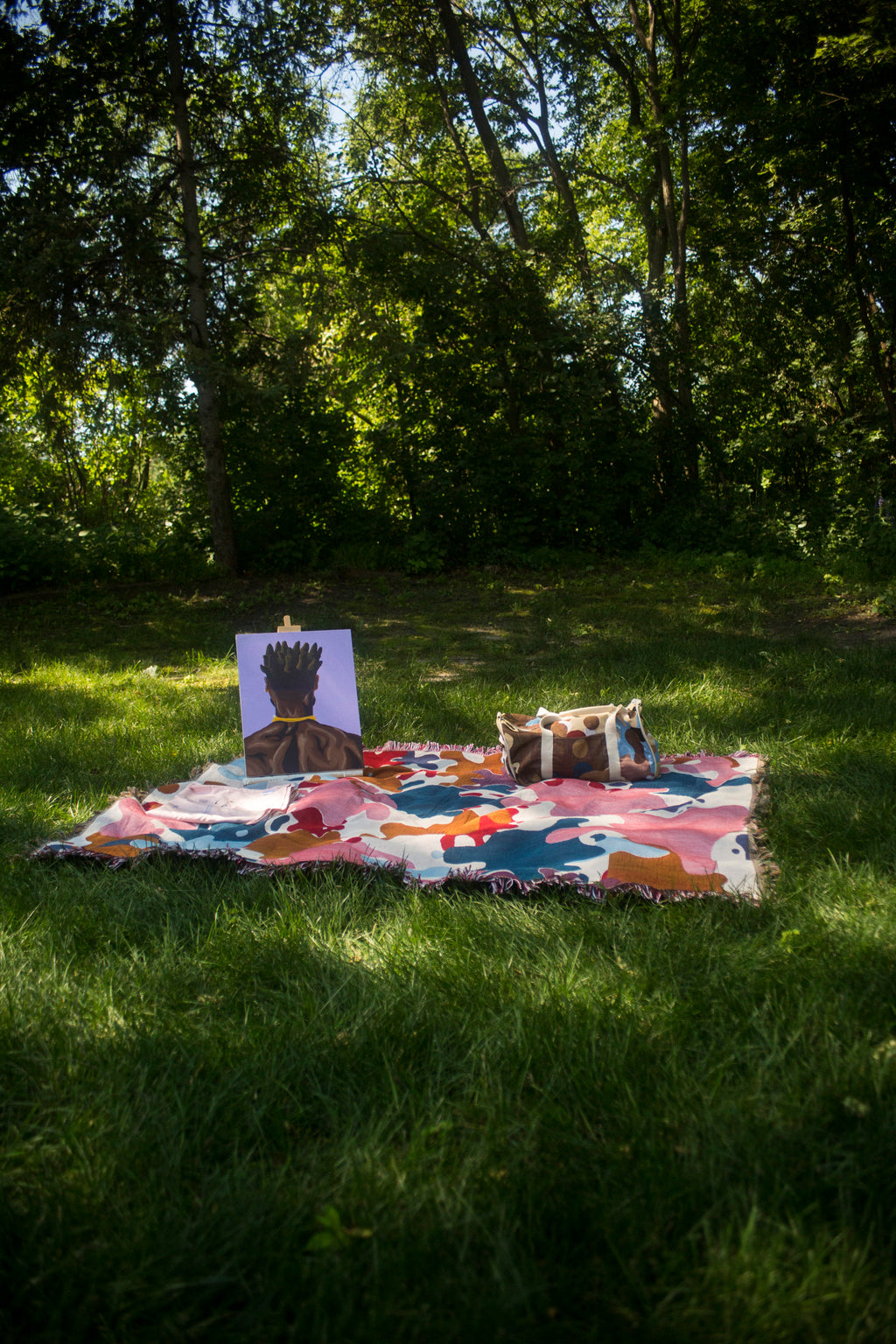 A colorful abstract patterned picnic blanket is spread on a grassy lawn in a wooded park setting. On the blanket sits a small canvas painting of a figure and a small bag.