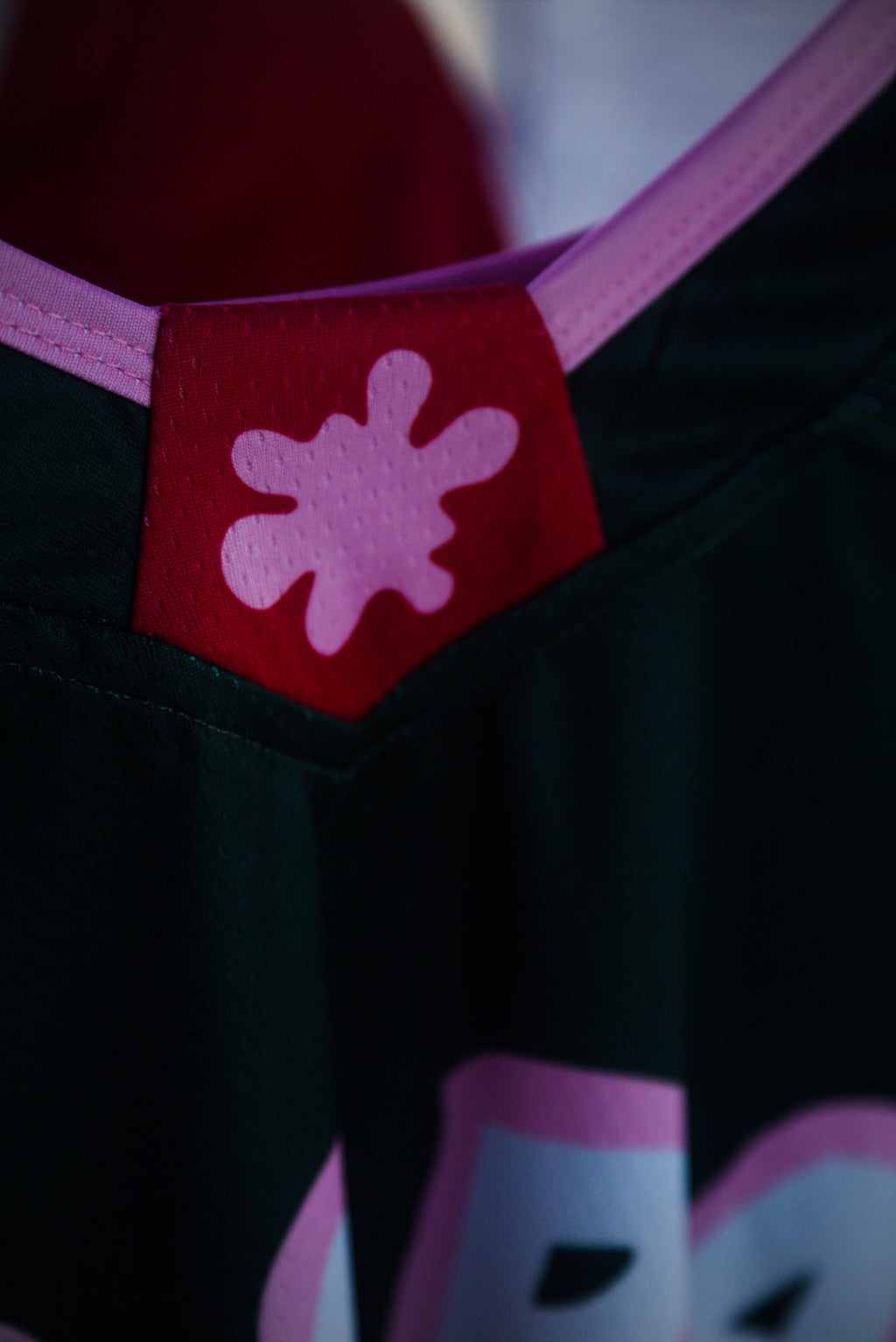 Close-up of a dark green athletic jersey with a red chest patch featuring a pink abstract splatter logo, and pink trim along the neckline.